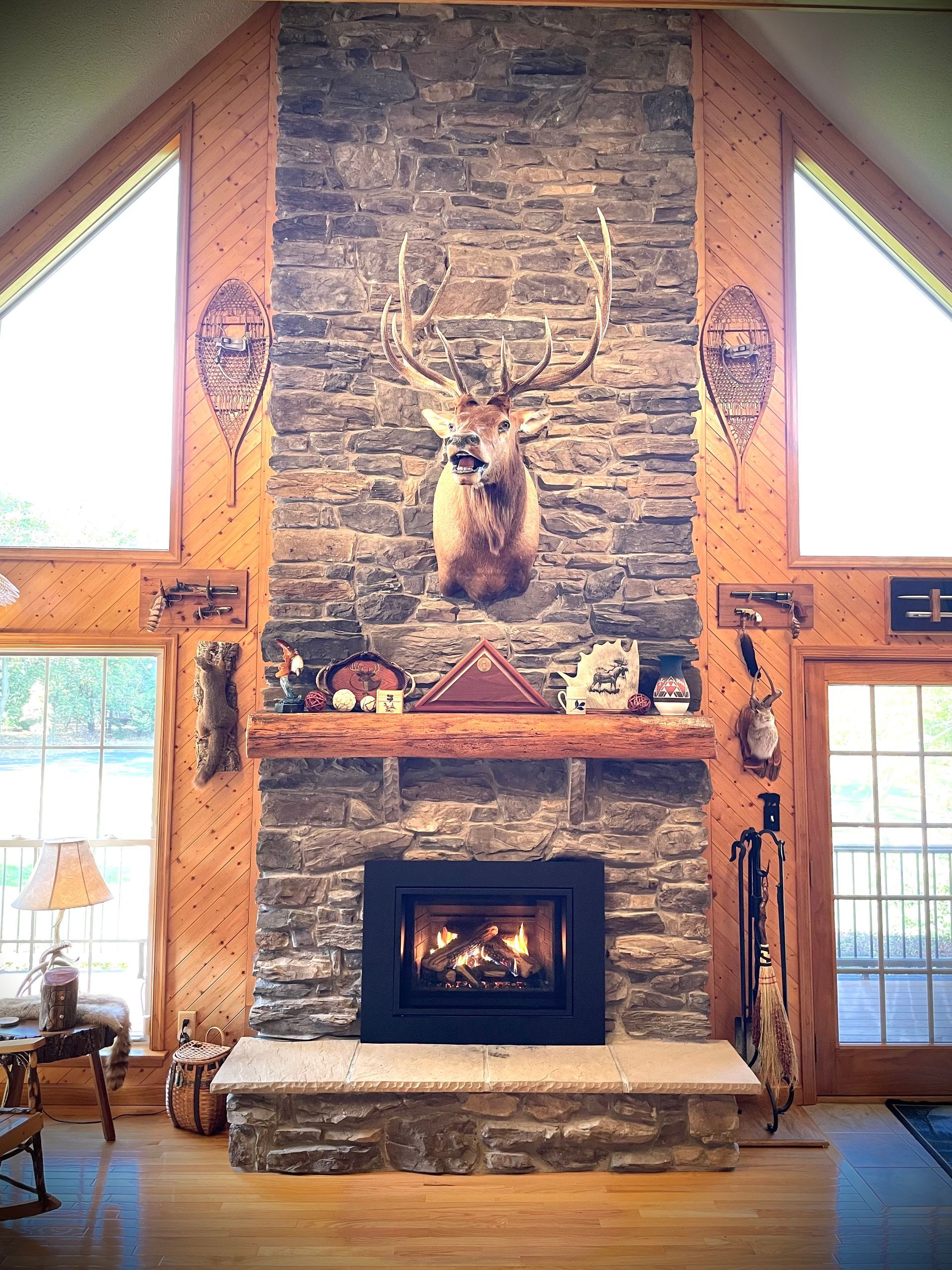 Lodge-style stone fireplace with mounted deer head and a lit fire in a wood-paneled great room