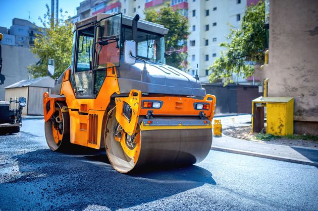 An orange and grey industrial road roller compacts freshly laid asphalt on a sunny city street.