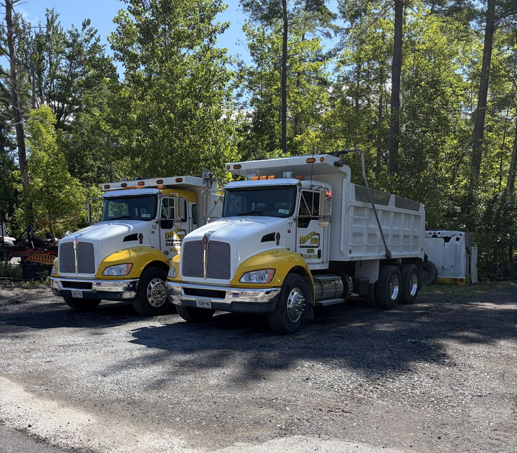 Two white dump trucks with yellow accents parked on gravel near a wooded area