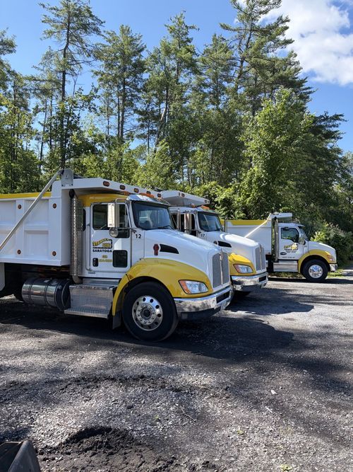 Two white dump trucks parked on gravel with trees and blue sky in the background