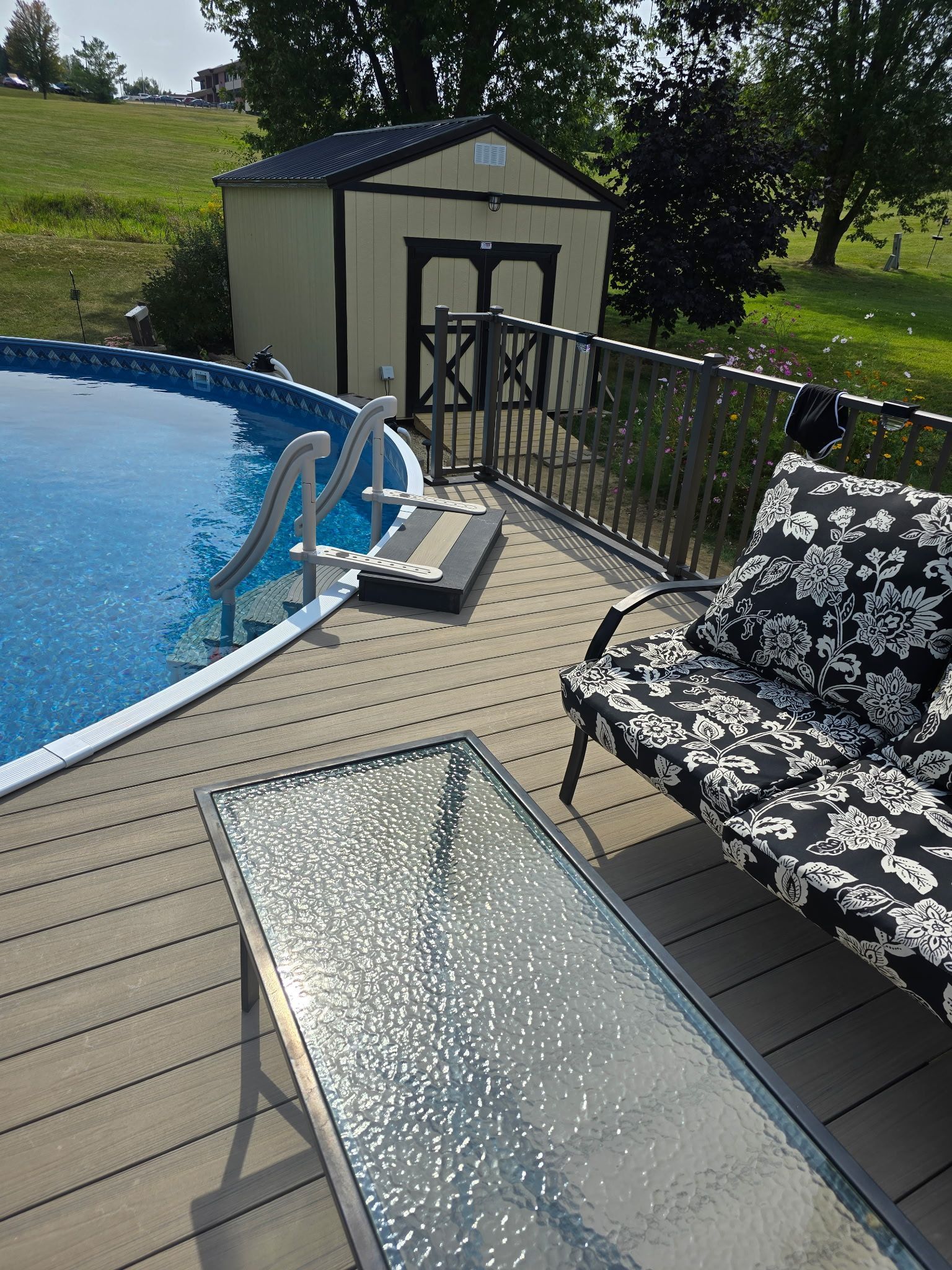 Backyard pool deck with glass railing, chair, and small shed in the background