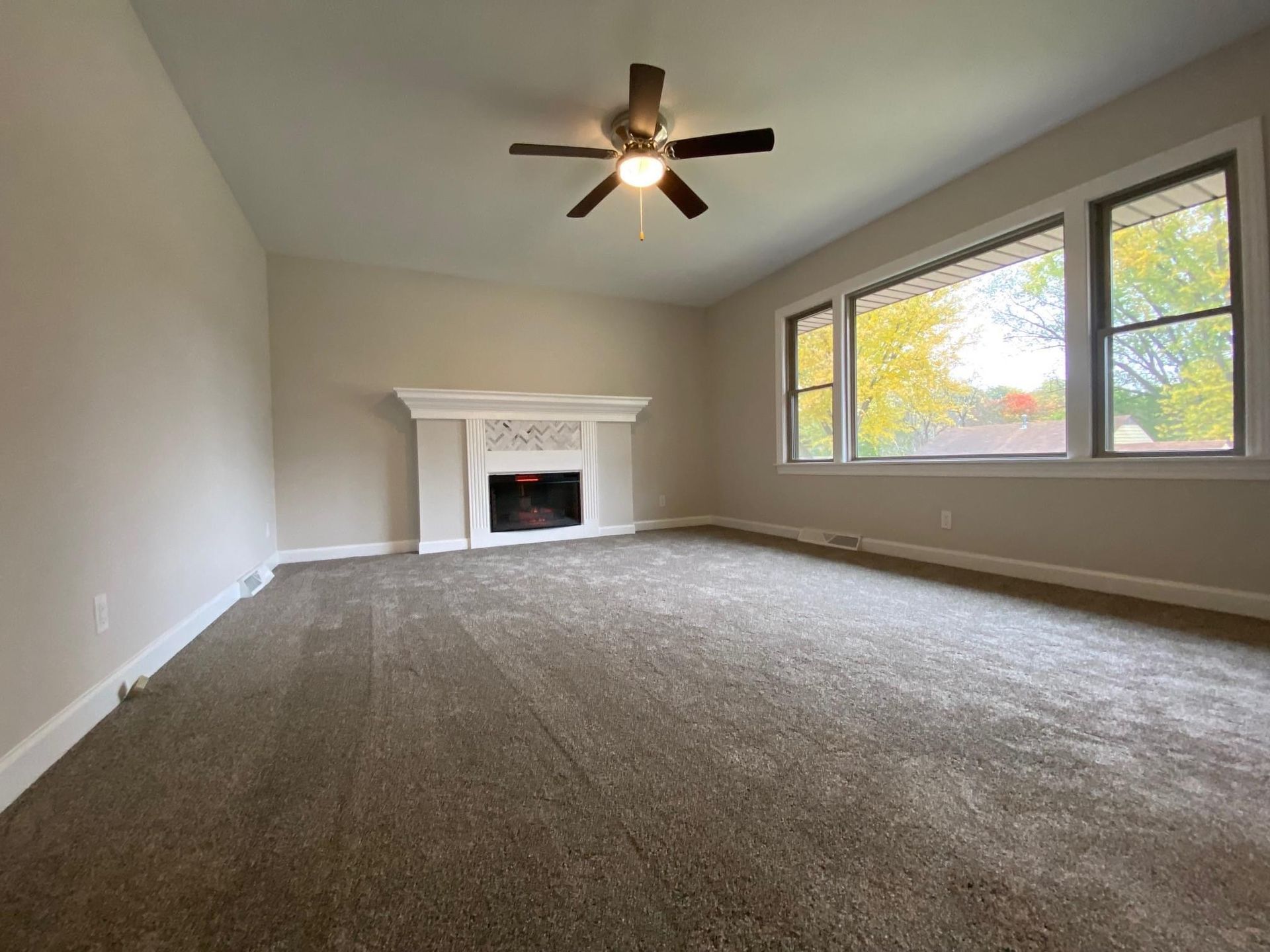 Empty carpeted living room with a white fireplace, ceiling fan, and large windows.