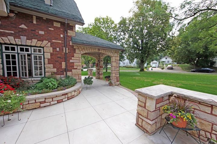 Brick house patio with white pavers, flower beds, and a low stone planter overlooking a green lawn.