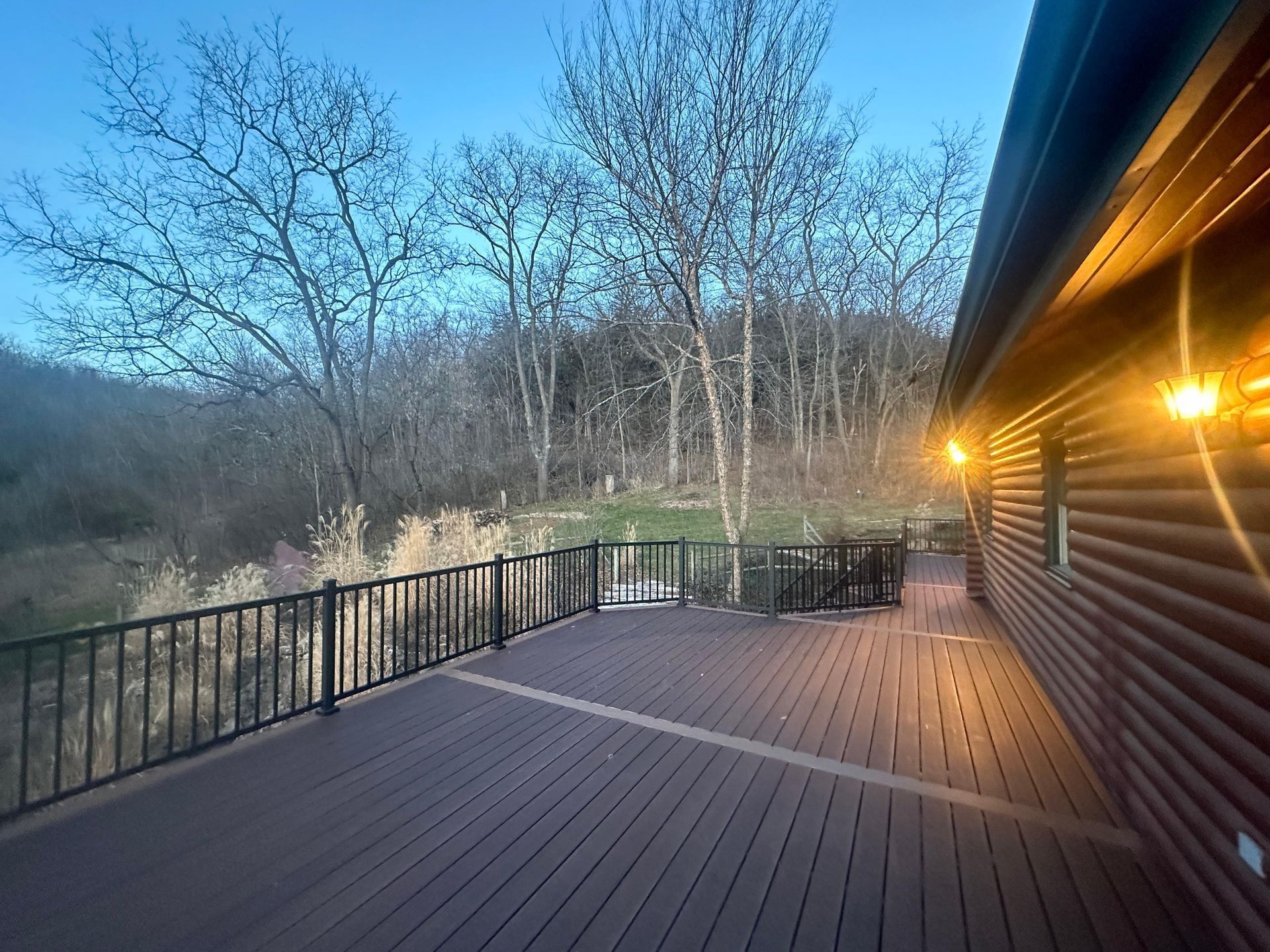 Deck beside log cabin at dusk, with railing, warm porch lights, and bare trees beyond.