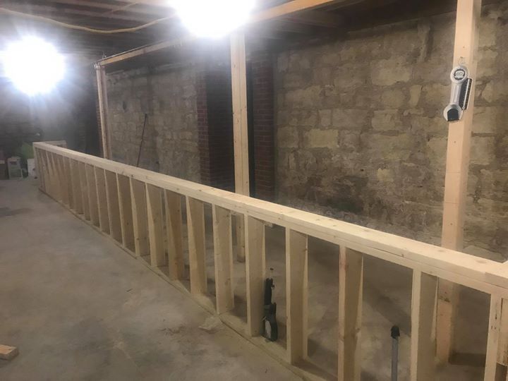 Wooden stair railing under construction in an unfinished basement, with exposed wood framing and cinderblock walls.