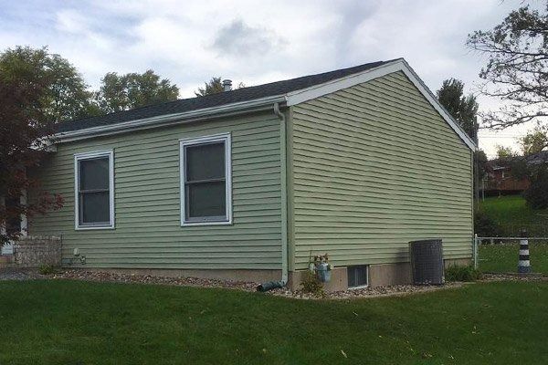 Single-story light green house with two windows and a sloped roof on a grassy lot.
