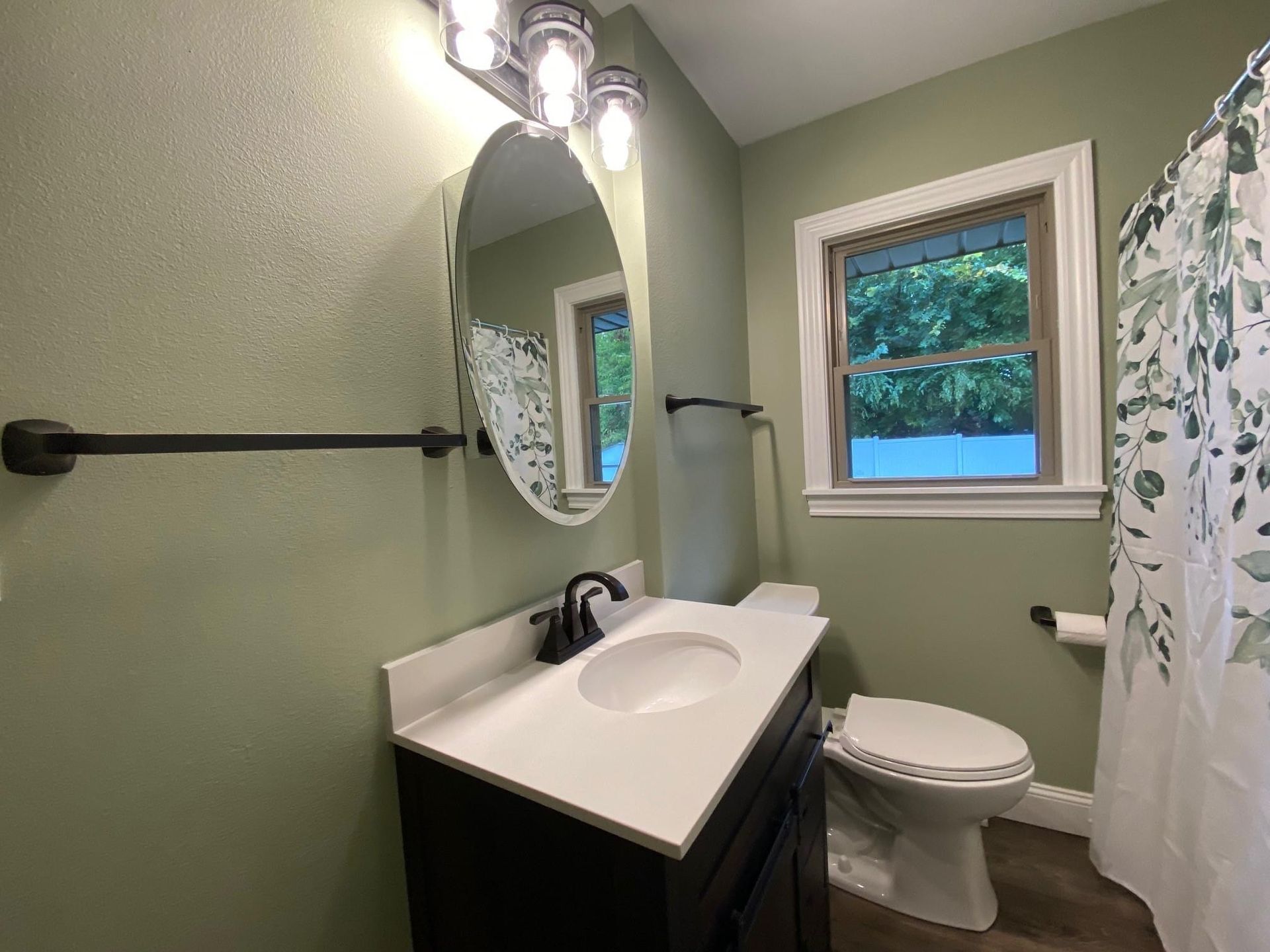 Small green bathroom with white sink vanity, toilet, oval mirror, and window.