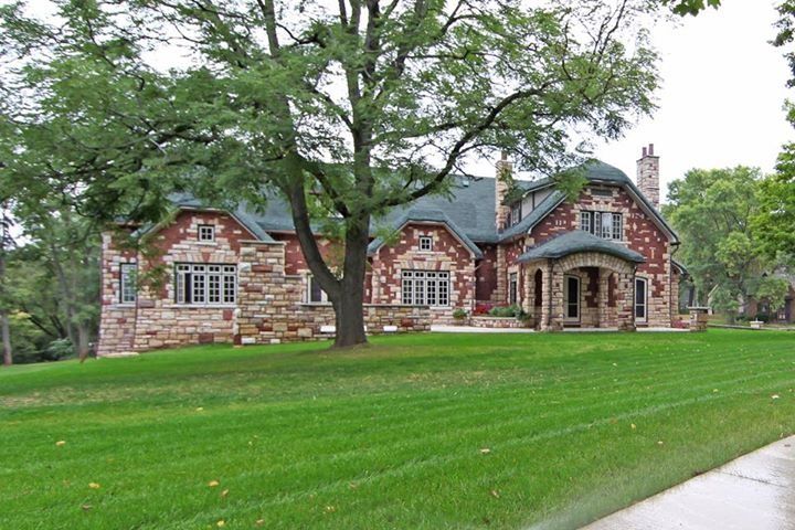 Stone house with arched porch and large tree, viewed across a green lawn