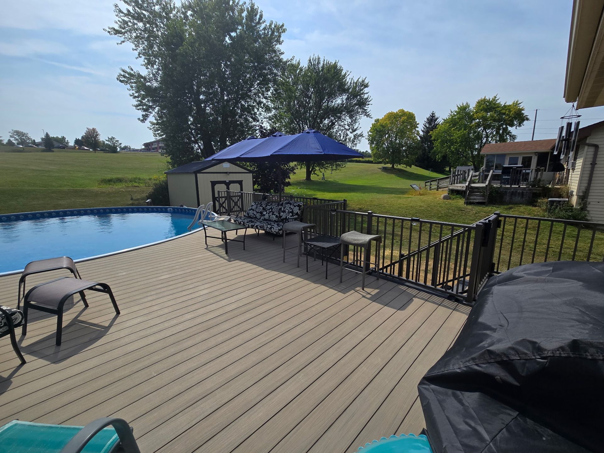 Patio deck beside a blue swimming pool, with black chairs, a grill, and a grassy yard by the water