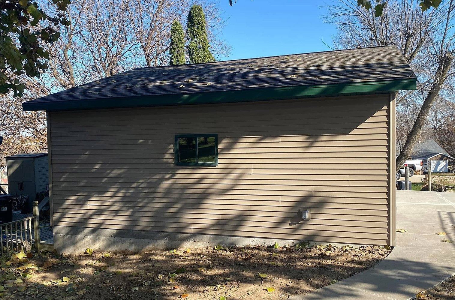 Side view of a tan house with a small window, shingled roof, and a driveway beside it under a blue sky.