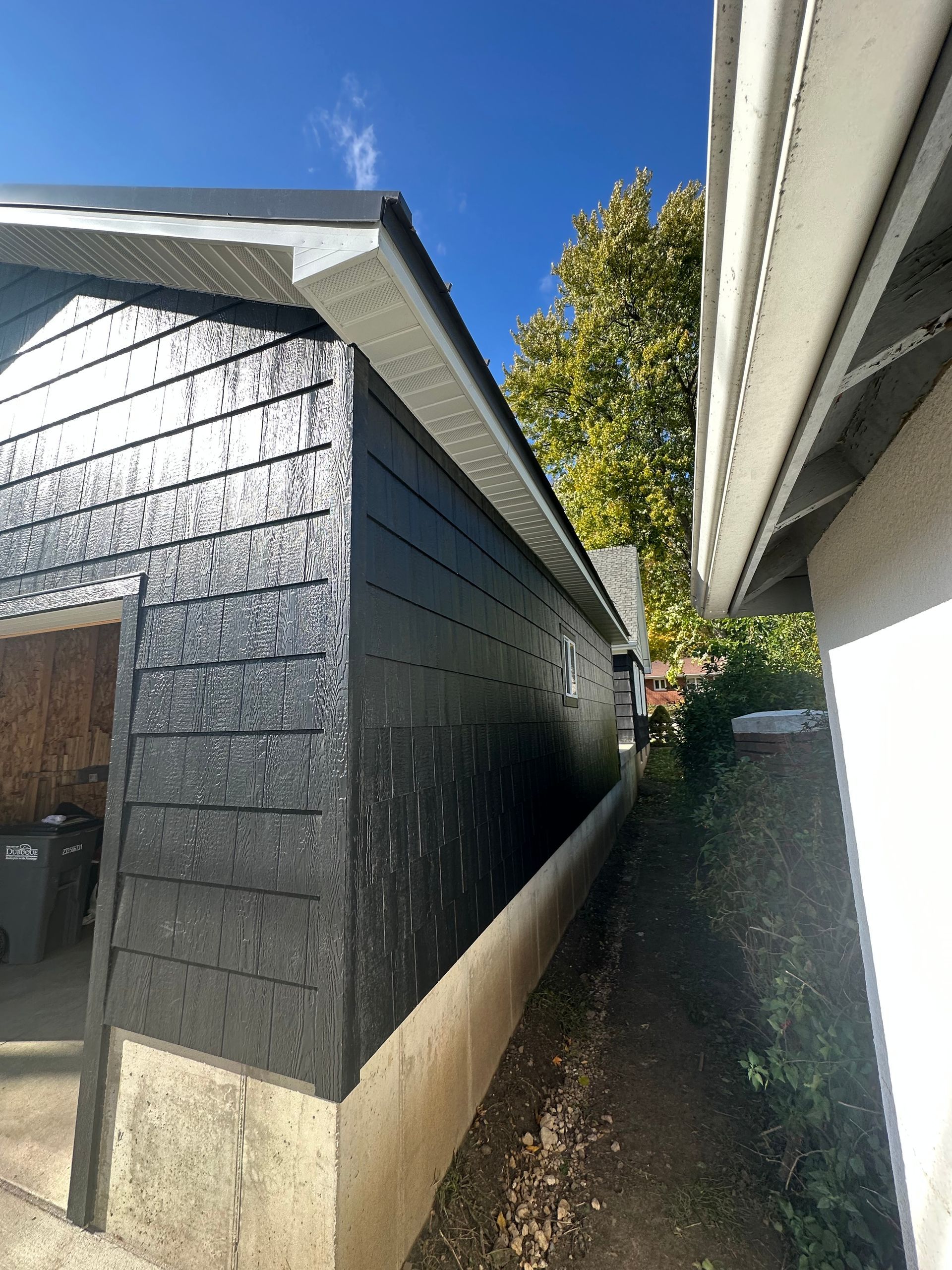 Narrow side passage between a black shed and white house under a blue sky