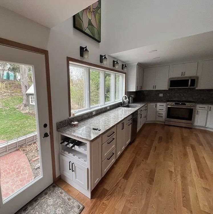 Bright modern kitchen with white cabinets, dark counters, wood floors, and large windows by a glass door