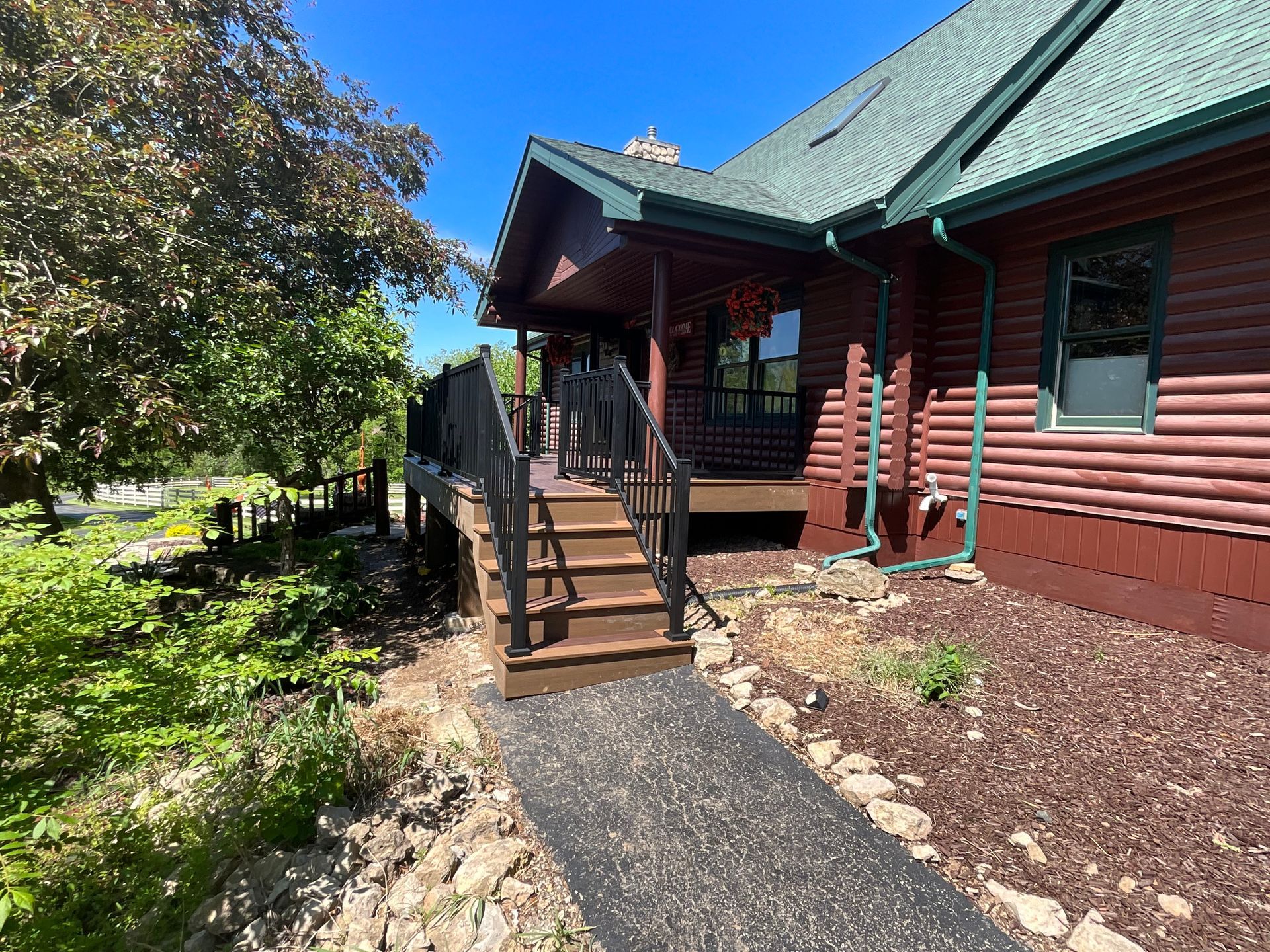 Log cabin entrance with wooden steps, railing, and a paved path beside landscaped mulch and trees