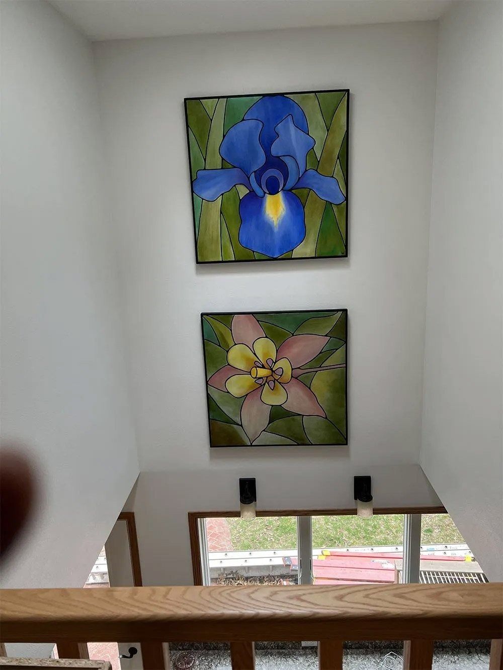Two flower paintings on a white stairwell wall above a window, one blue and one pink-yellow.