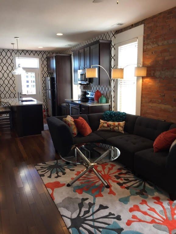 Cozy living room with dark sofa, glass coffee table, patterned rug, and kitchen area in the background