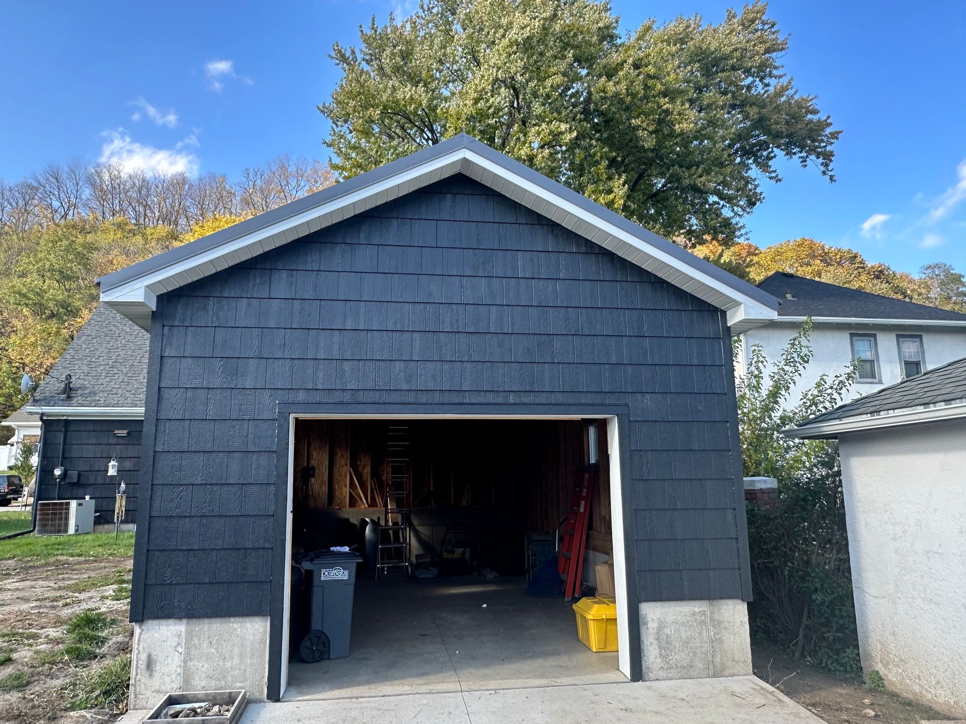 Detached black garage with open door, set against a house and trees under a blue sky