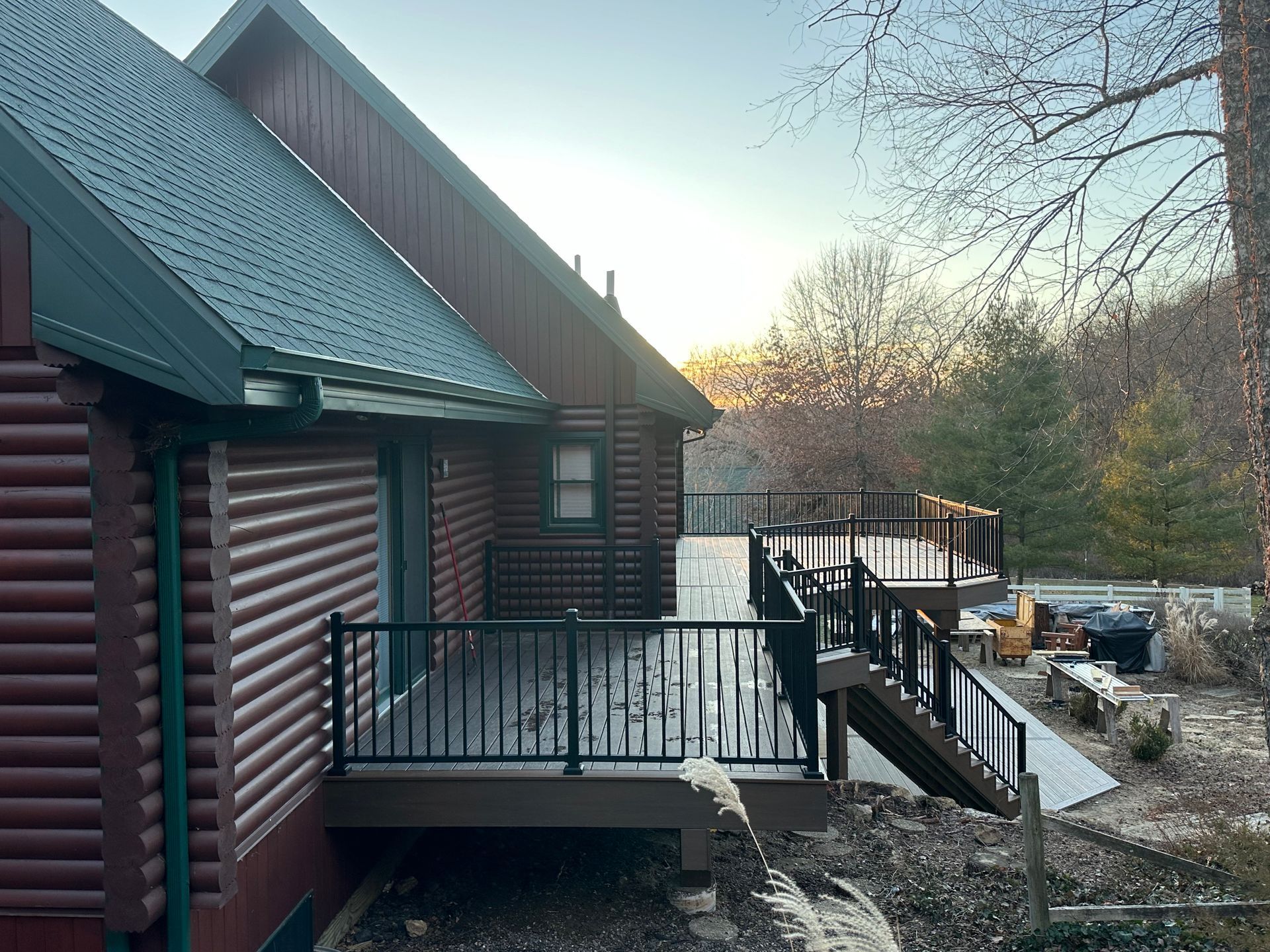 Log cabin deck with black railings and stairs, dusted with snow beside a wooded yard.
