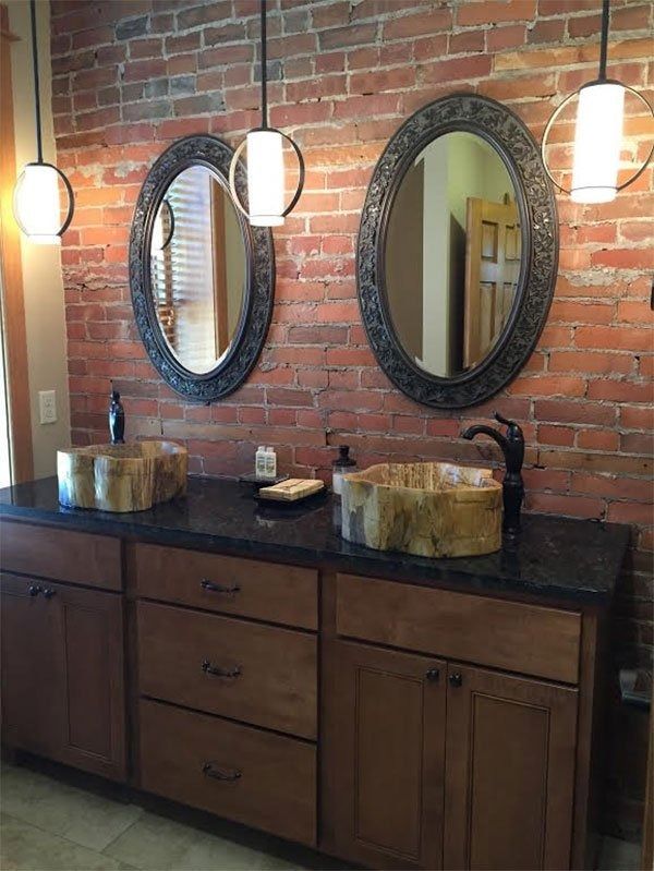 Bathroom vanity with dark wood cabinets, two oval mirrors, hanging lights, and brick wall backdrop