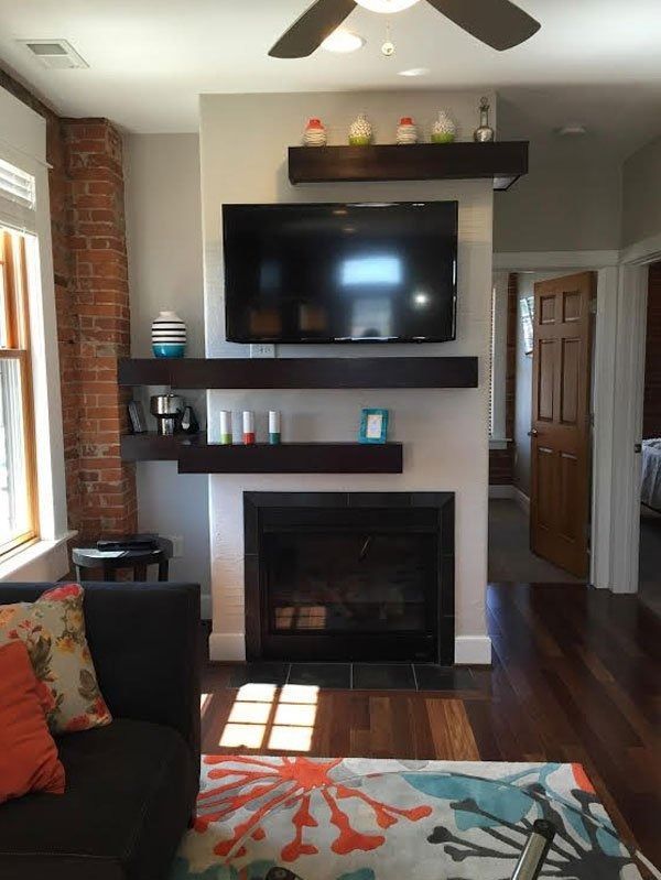 Living room with wall-mounted TV above fireplace, dark wood floors, shelves, and colorful rug