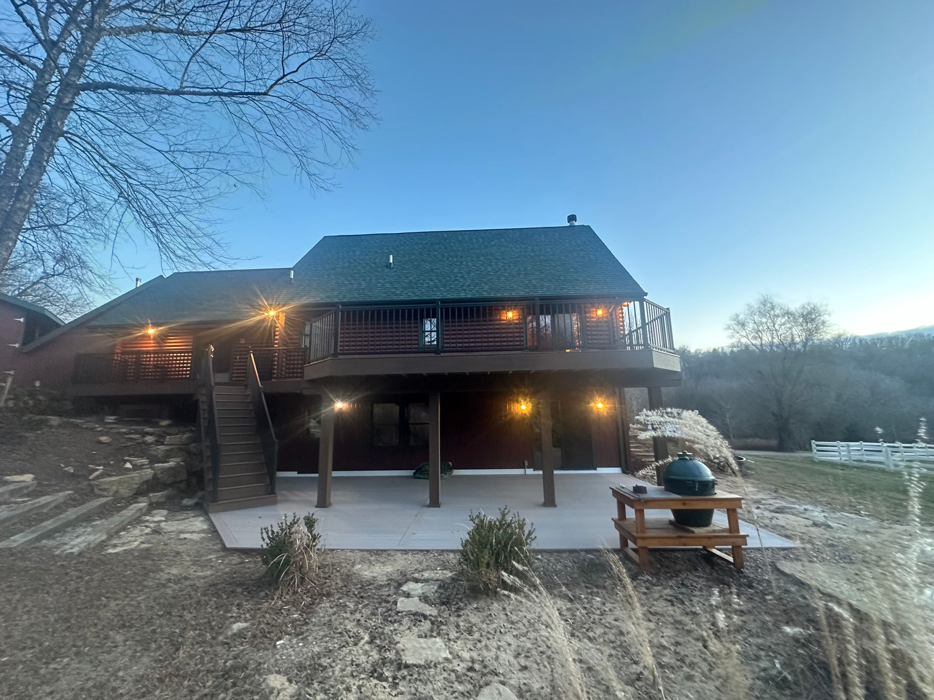 Rustic cabin with wraparound porch at dusk, lit windows, and a snowy yard with a wooden table and bench
