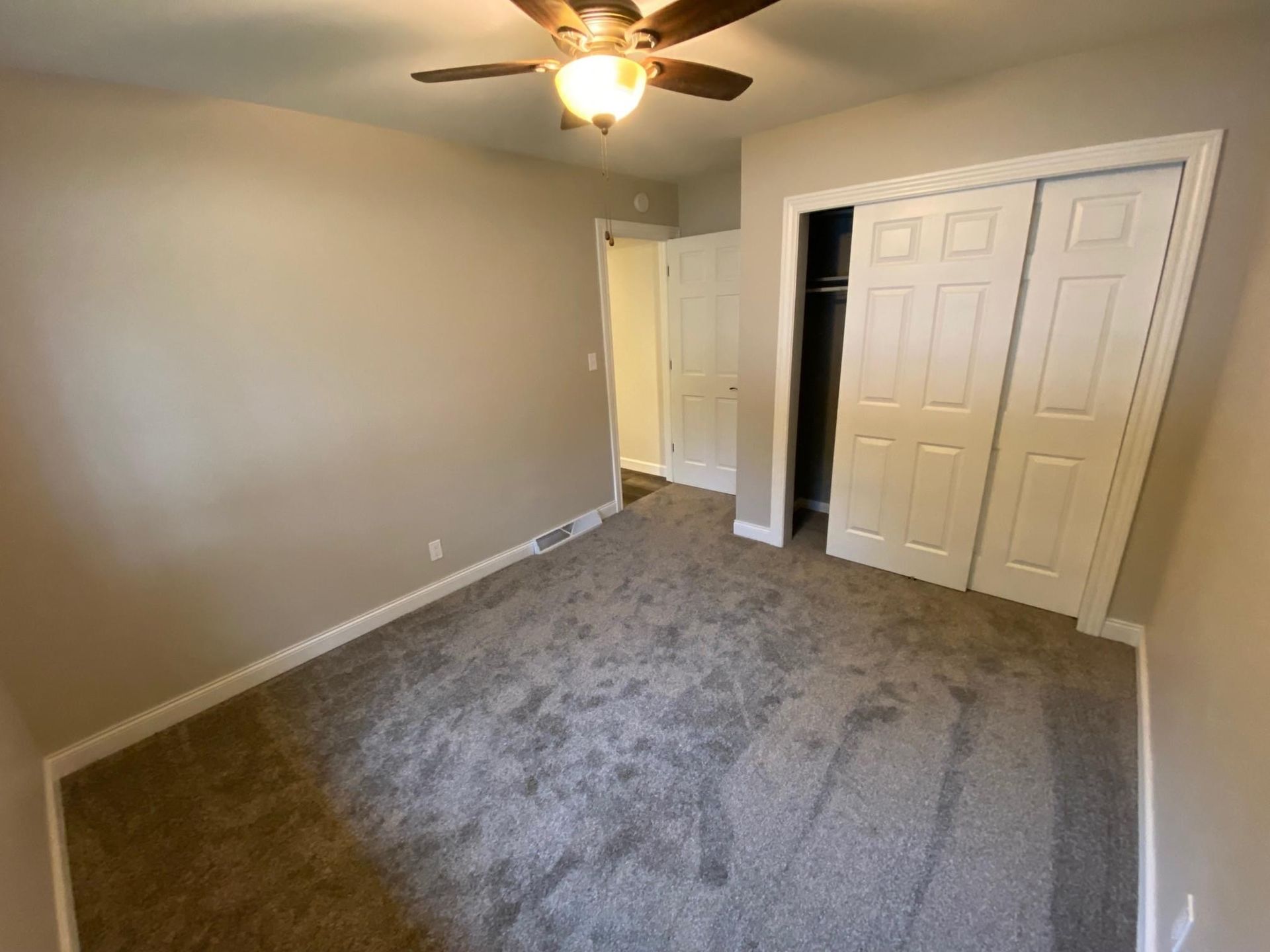 Empty beige bedroom with gray carpet, ceiling fan, open doorway, and sliding closet doors.
