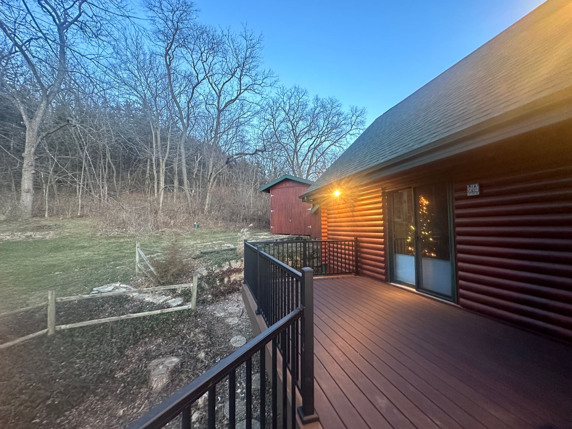 Wooden cabin deck with black railing at dusk, lit porch light, overlooking a wooded yard