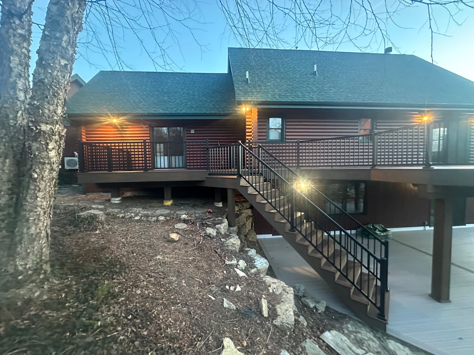 Log cabin with lit porch and exterior staircase on a wooded hillside at dusk