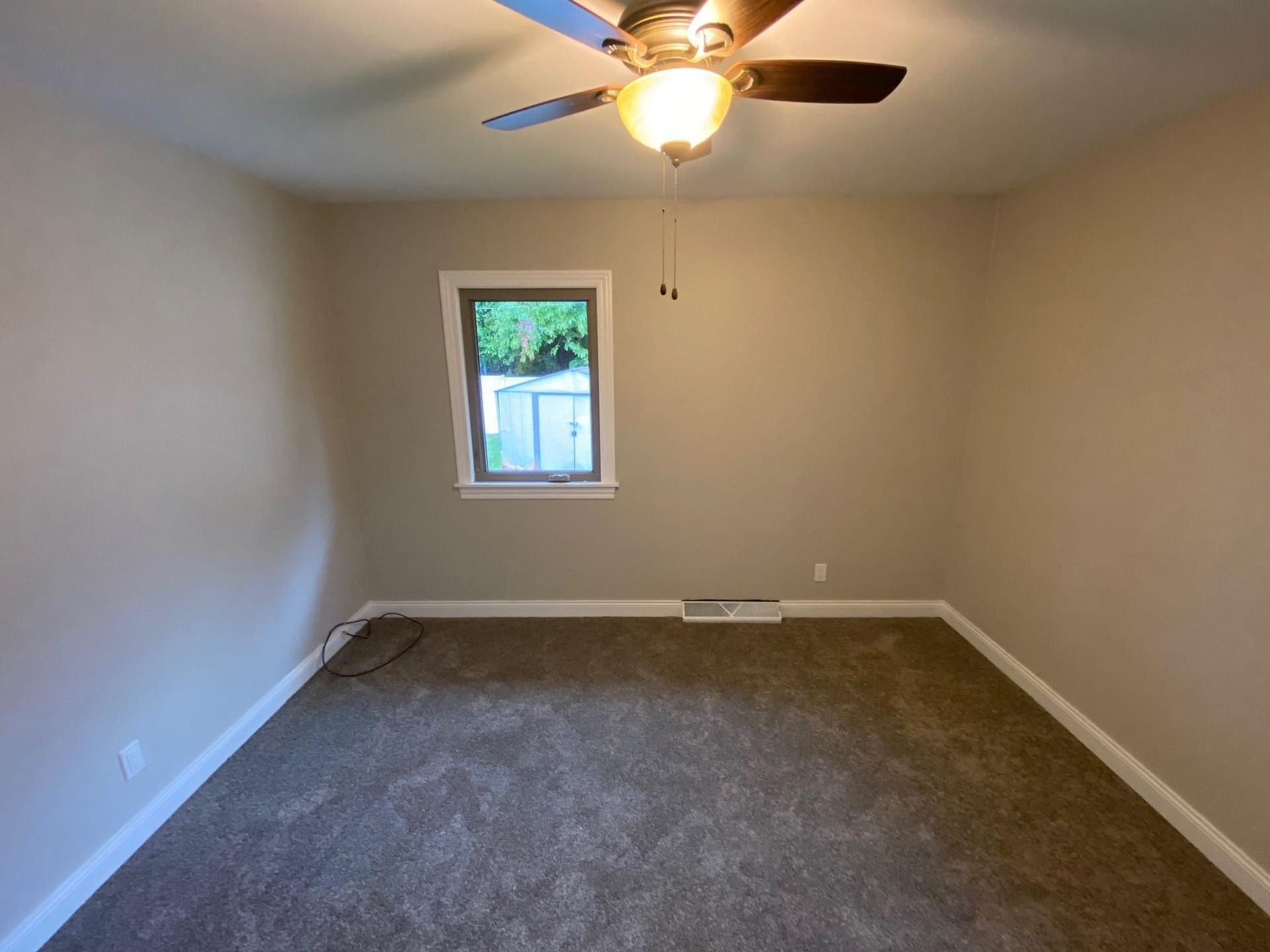 Empty beige bedroom with carpet, ceiling fan, and a small window showing greenery