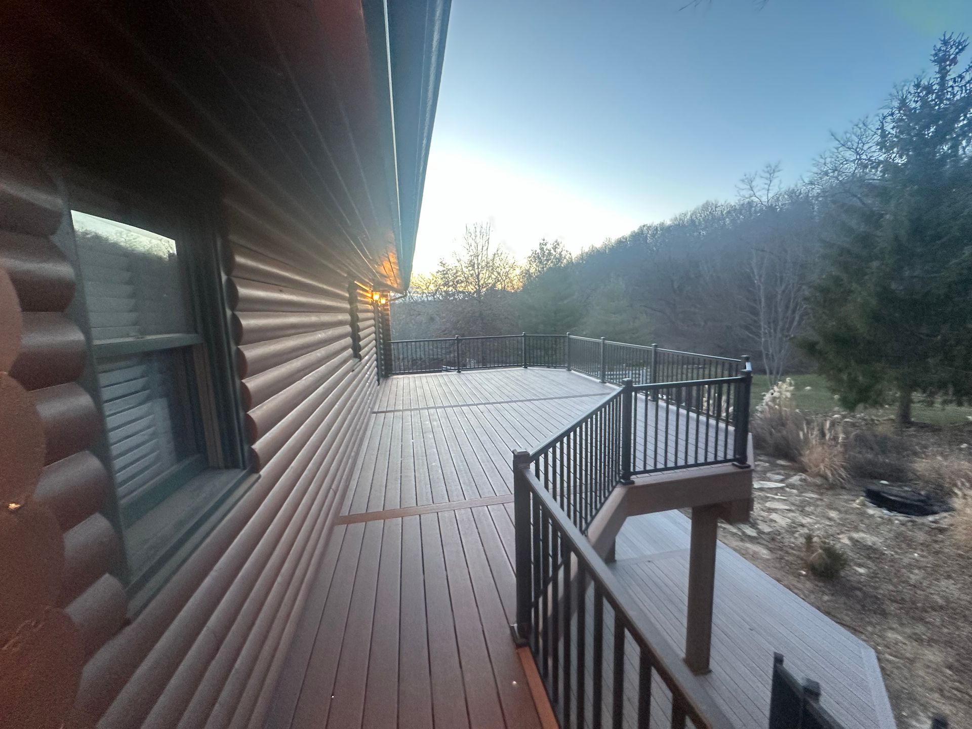 Wooden deck beside a log cabin at sunrise, with black railings and a wooded yard beyond.