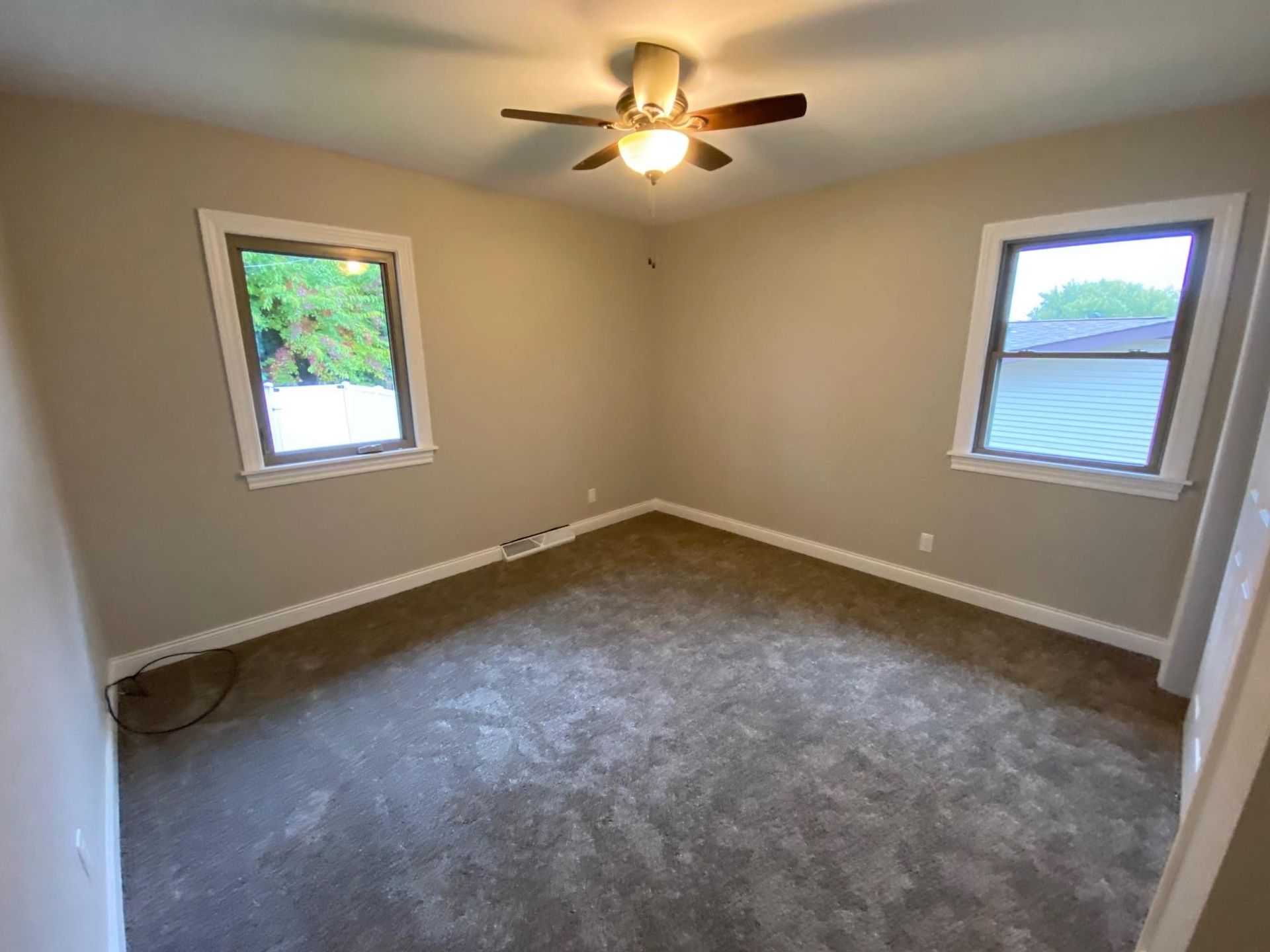 Empty beige bedroom with carpet, two windows, and a ceiling fan with lights