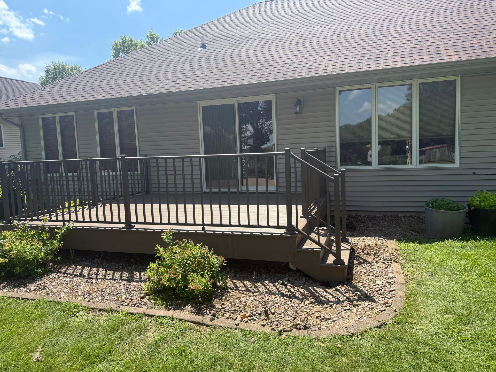 Back porch with black railings, tan siding, and large windows, bordered by landscaped rocks and grass.