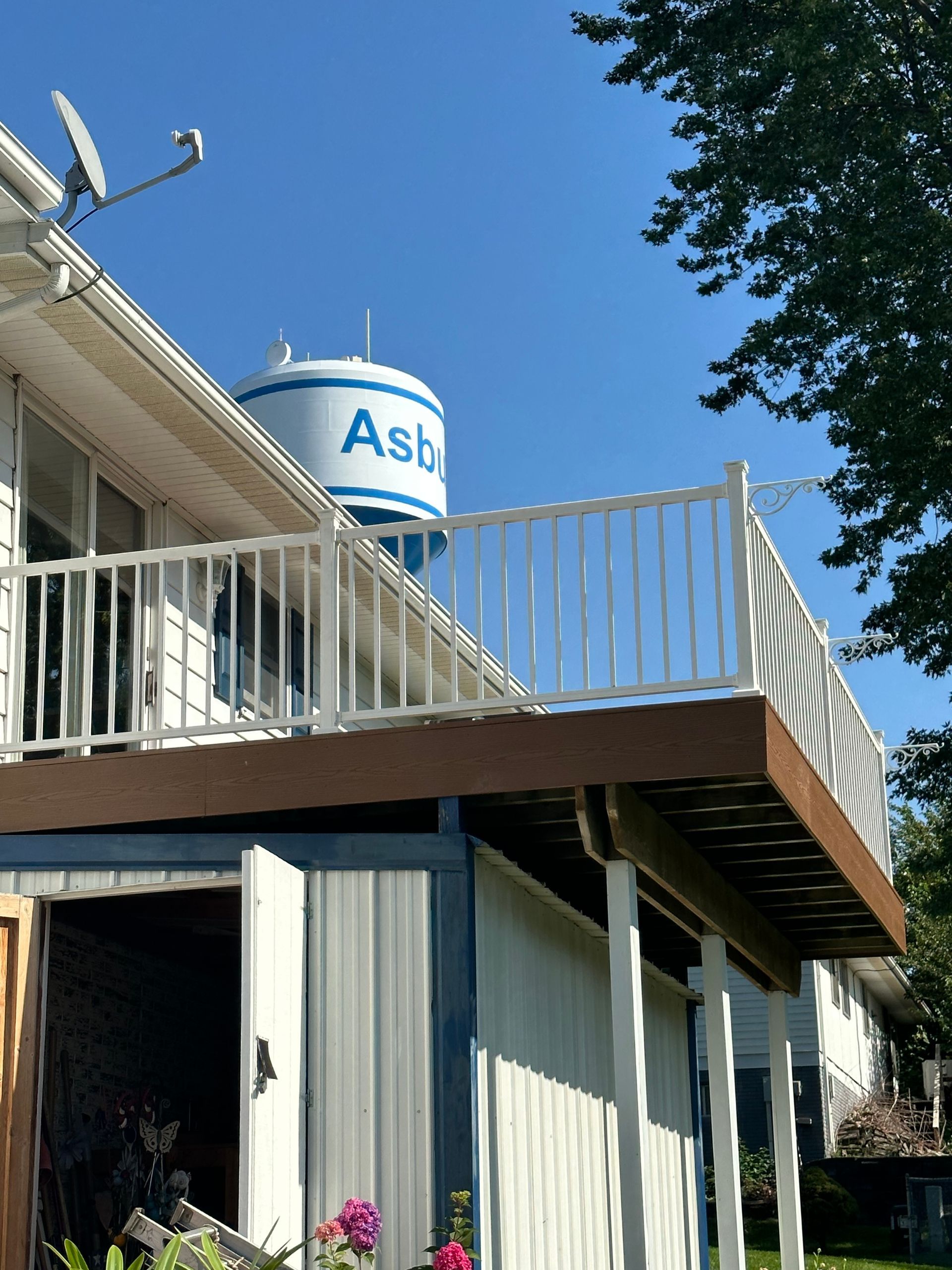 Blue water tower labeled “Ast” above a house deck and white railing under a clear sky.