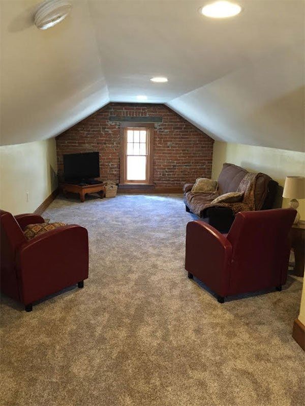 Small attic living room with sloped ceiling, brick wall, window, carpet, and red chairs with a sofa.