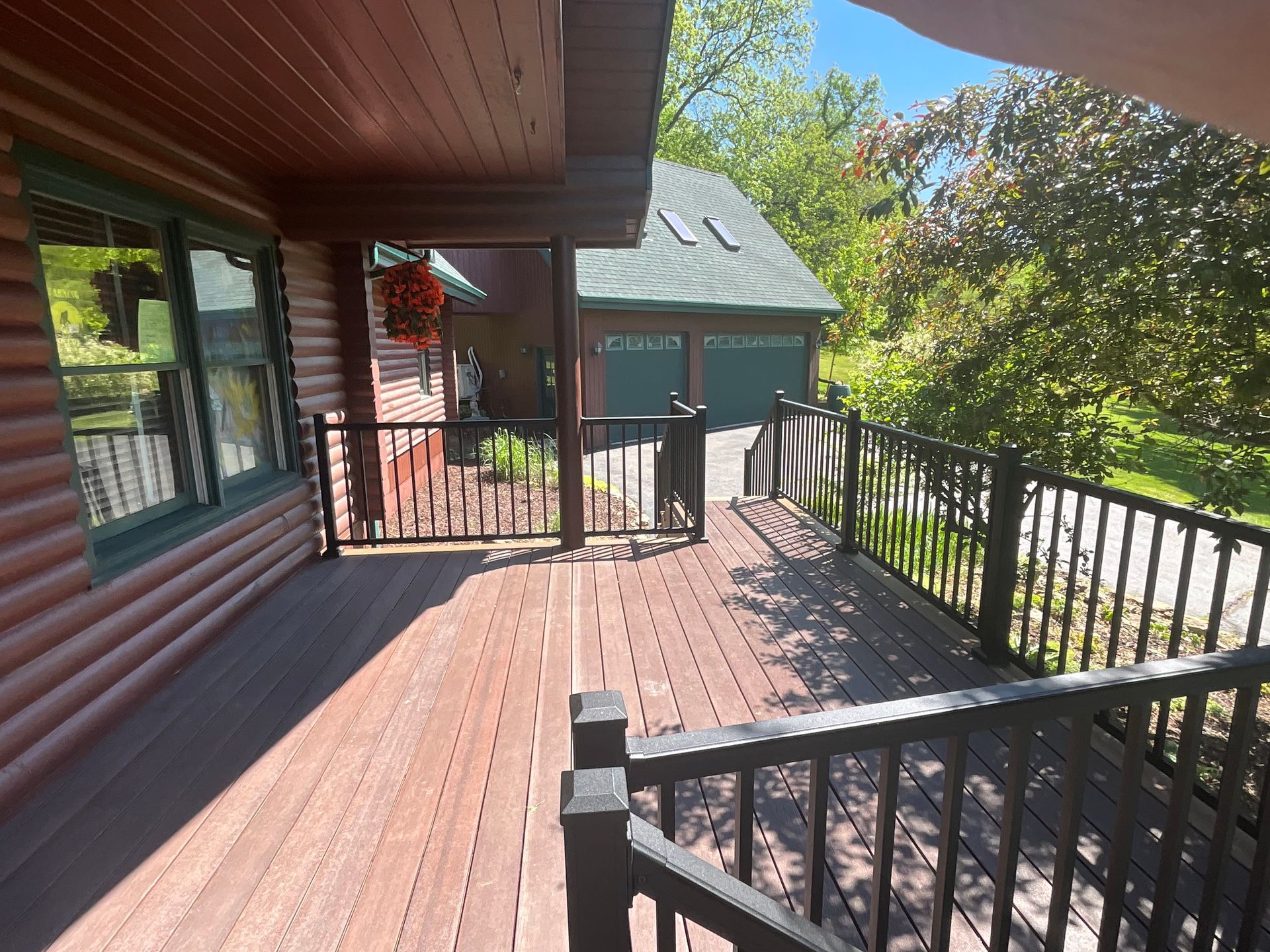 Wooden deck with black railings beside a log cabin, overlooking trees and a green shed in sunlight
