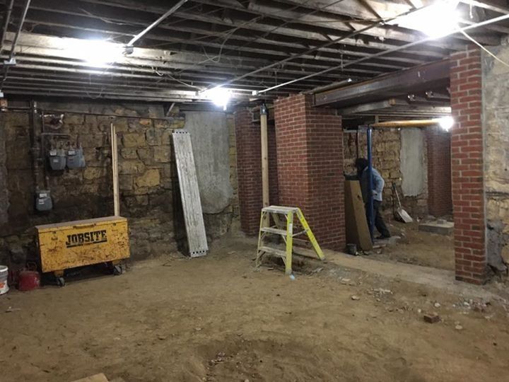 Basement under renovation with exposed beams, brick columns, tools, and a worker in the background