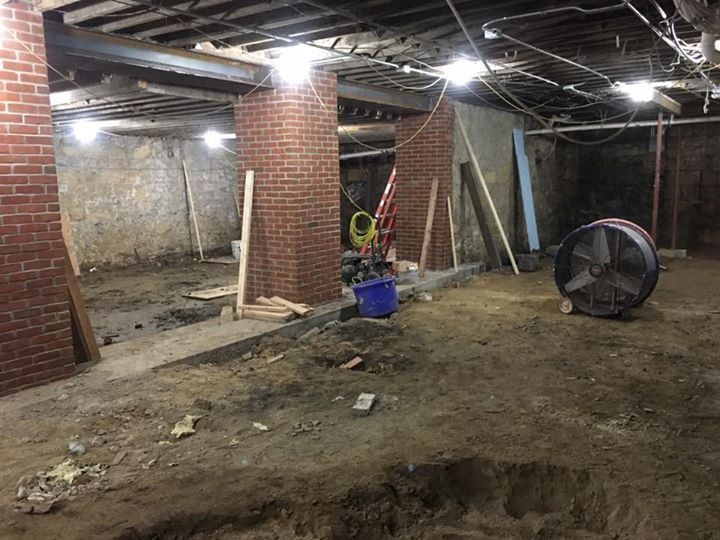 Unfinished basement with exposed beams, brick pillars, scattered tools, and a large floor hole.