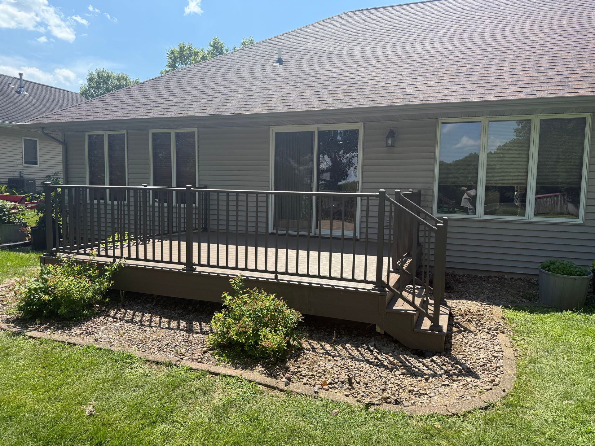 Back deck with black railing on a beige house, set in a grassy yard under a bright sky