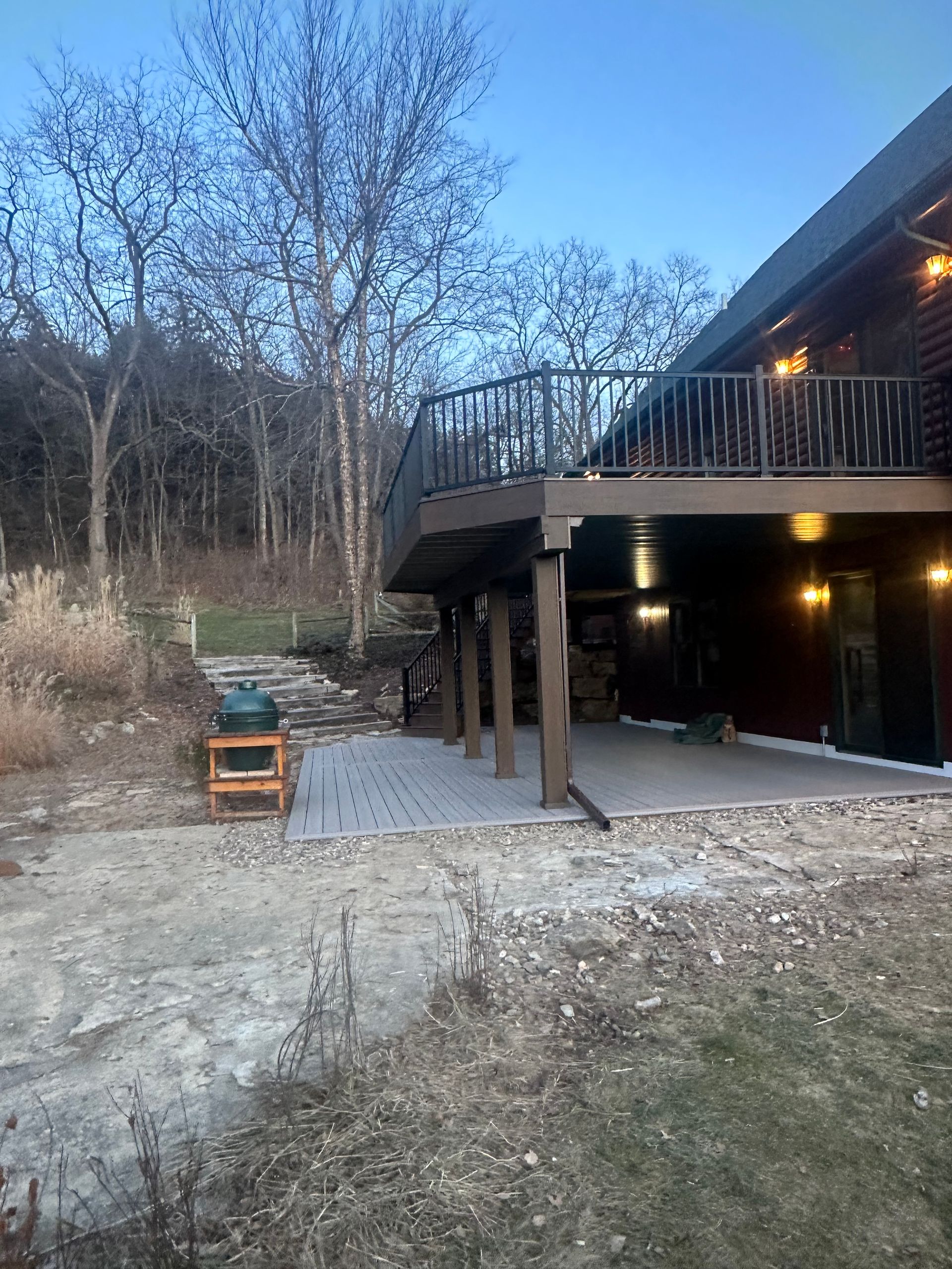 Backyard deck with stairs, warm lights, and a fire pit beside leafless trees at dusk