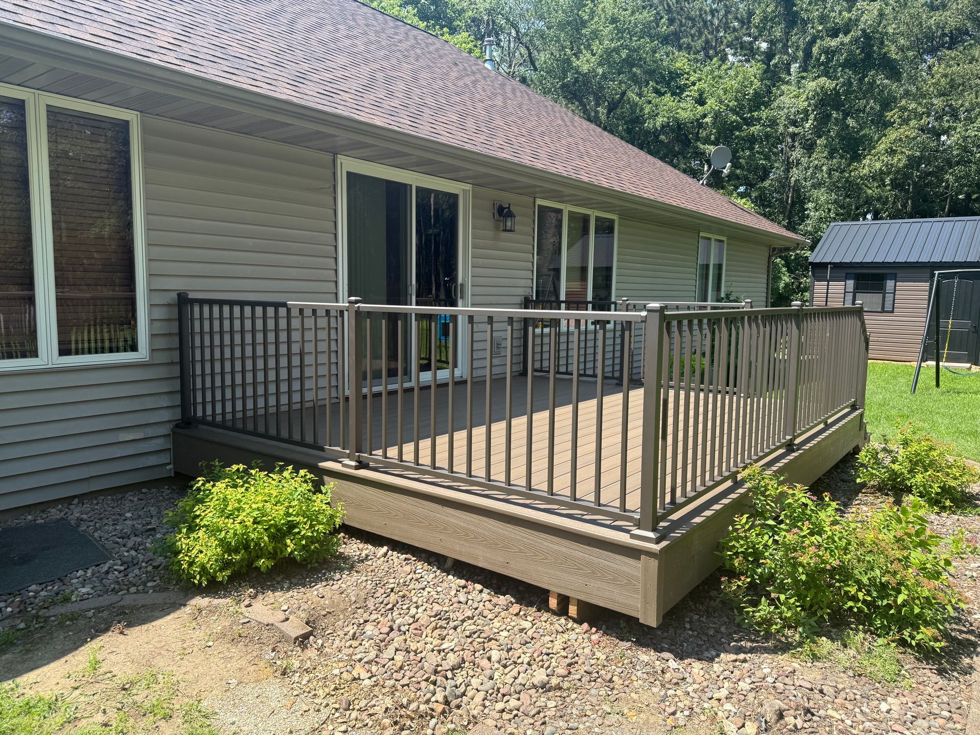 Raised wooden deck with metal railing attached to a house beside a gravel yard and shrubs