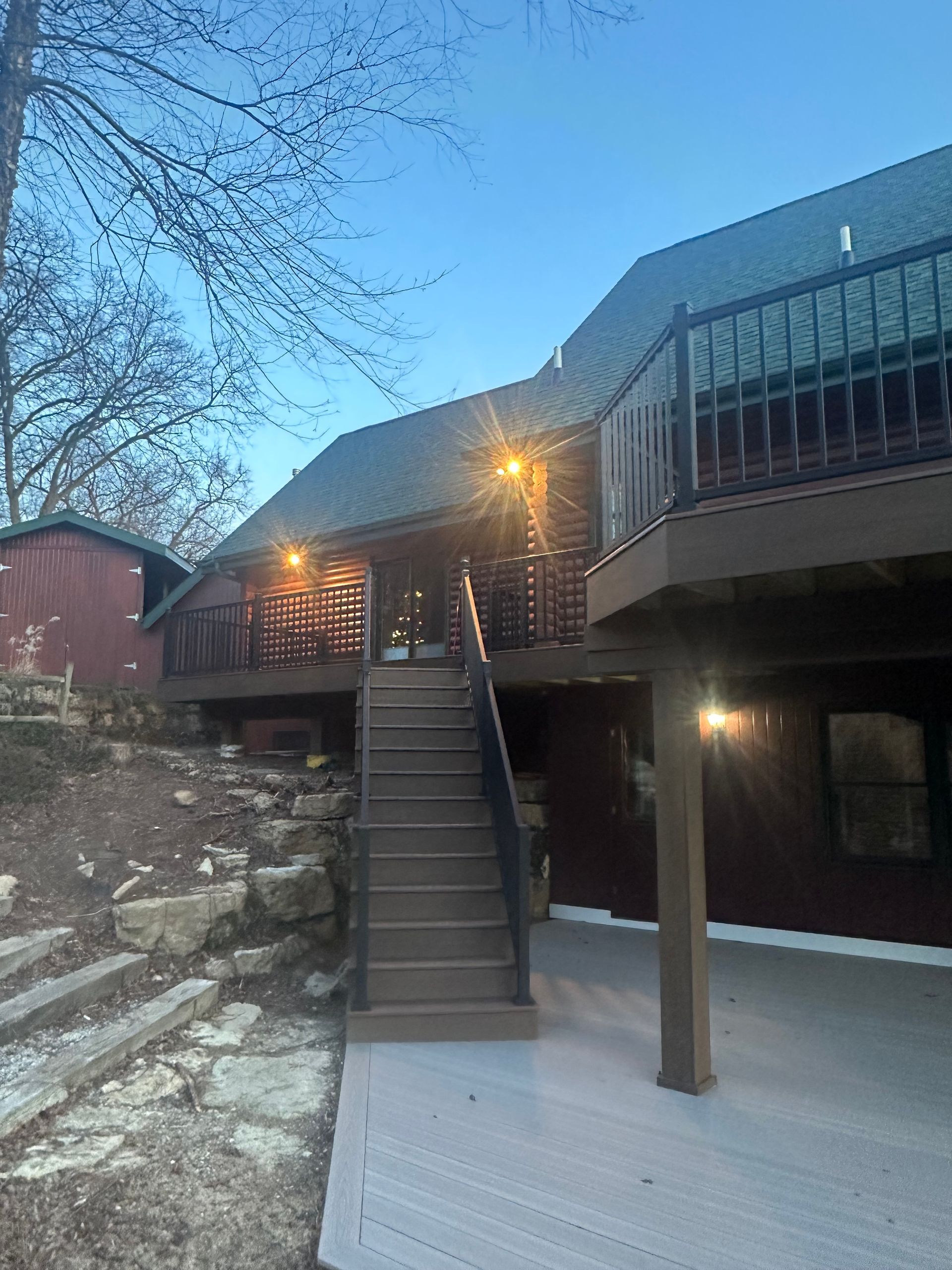 Snowy house exterior with wooden deck, stairs, and warm lights at dusk