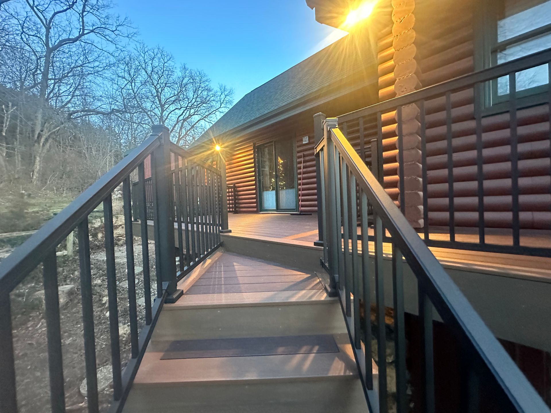Wooden ramp leading to a log cabin entrance at sunset, with warm lights and bare trees nearby