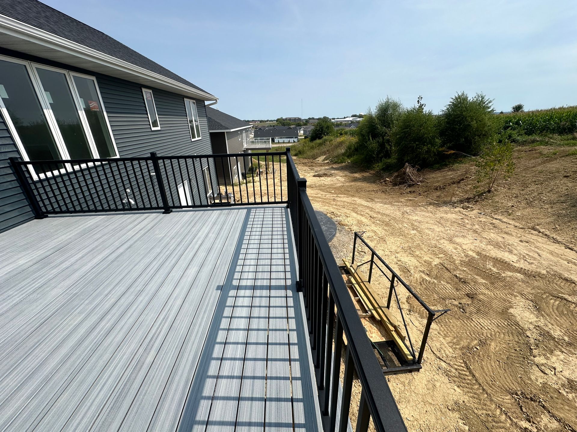 Deck with black railing beside a sandy slope and greenery under a clear sky