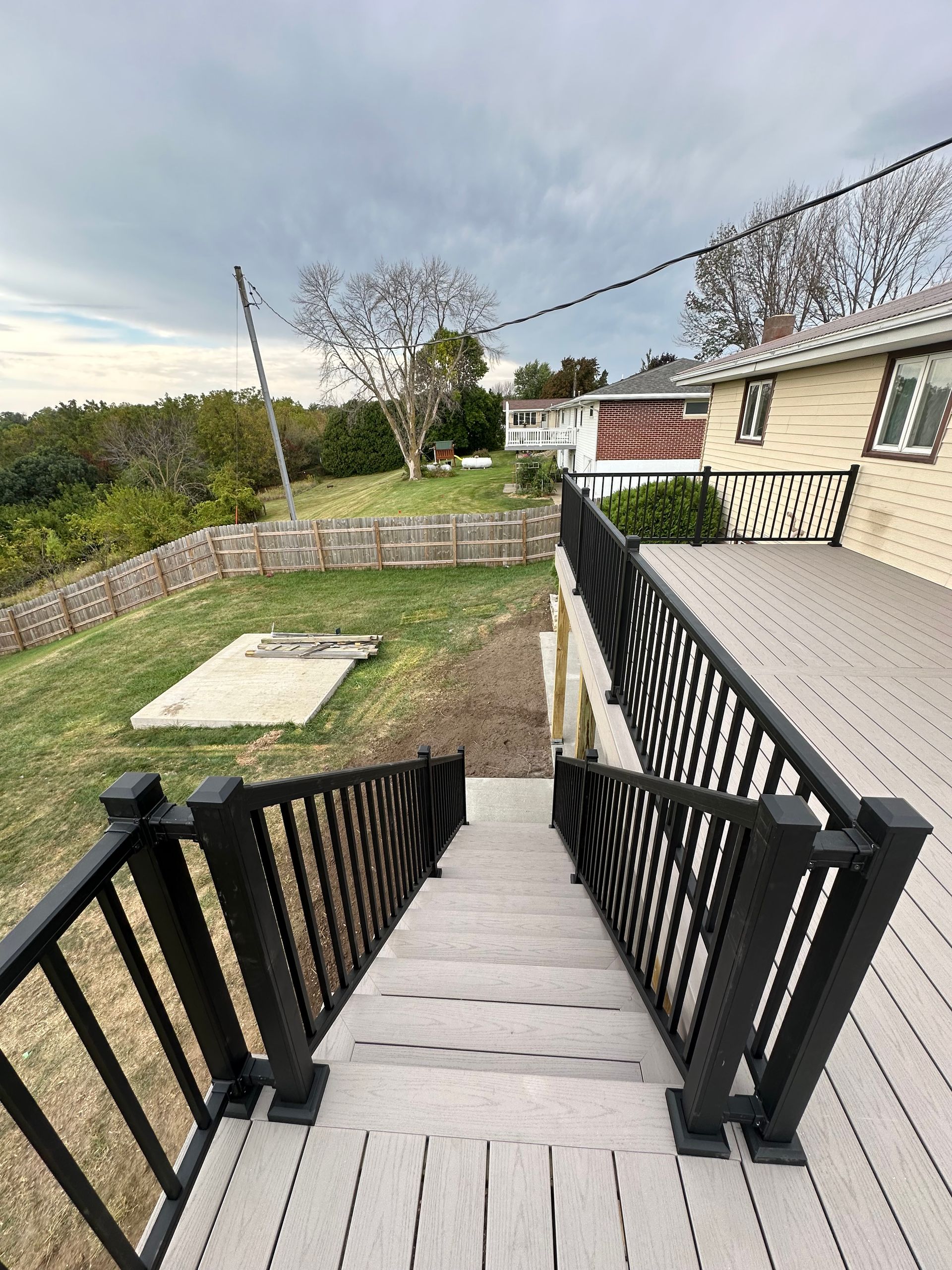 Outdoor stairs with black railings descending to a grassy yard beside a beige house and stone wall.