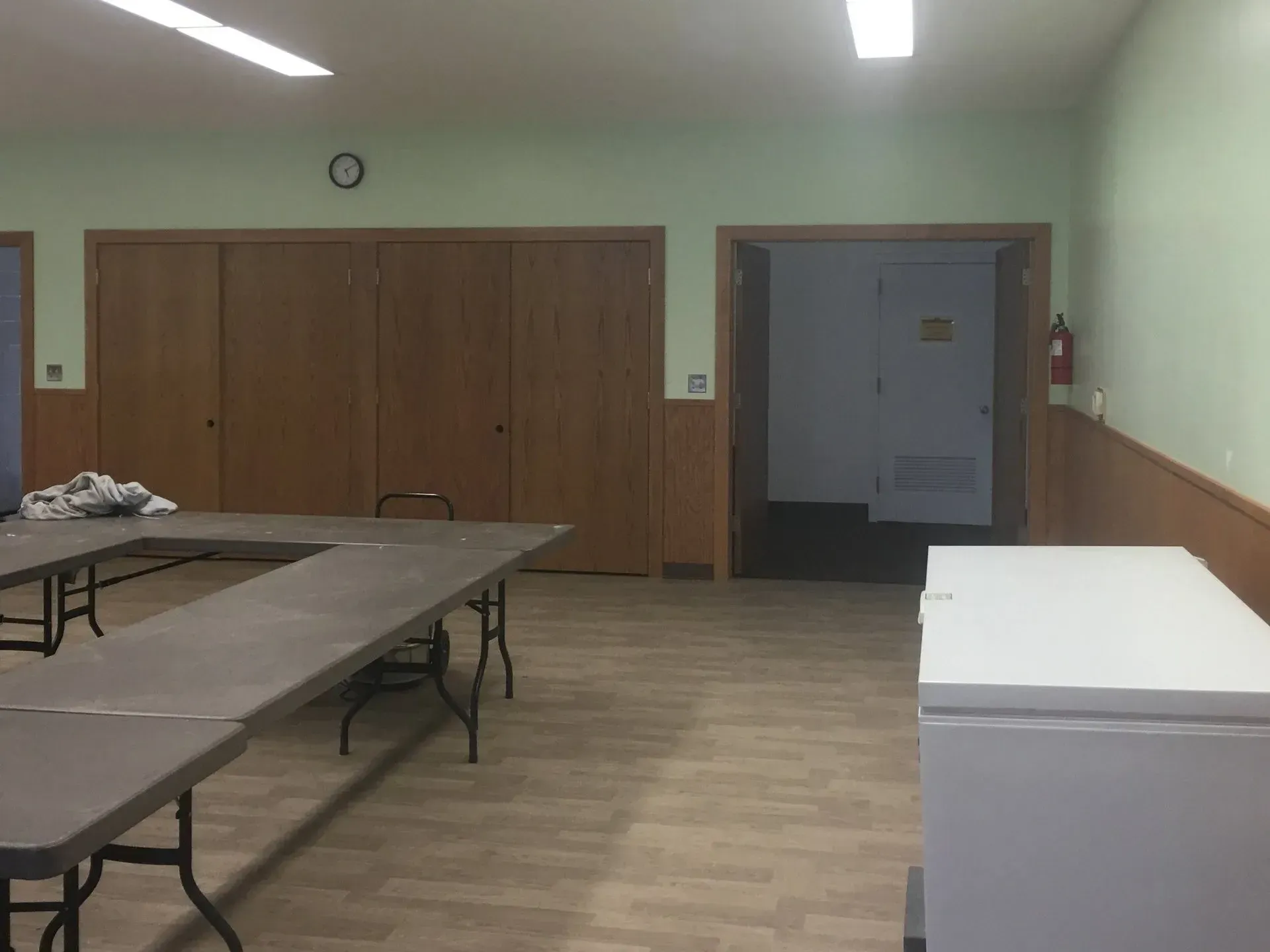 Empty room with tables, folding chairs, wood-paneled walls, and open doorway.