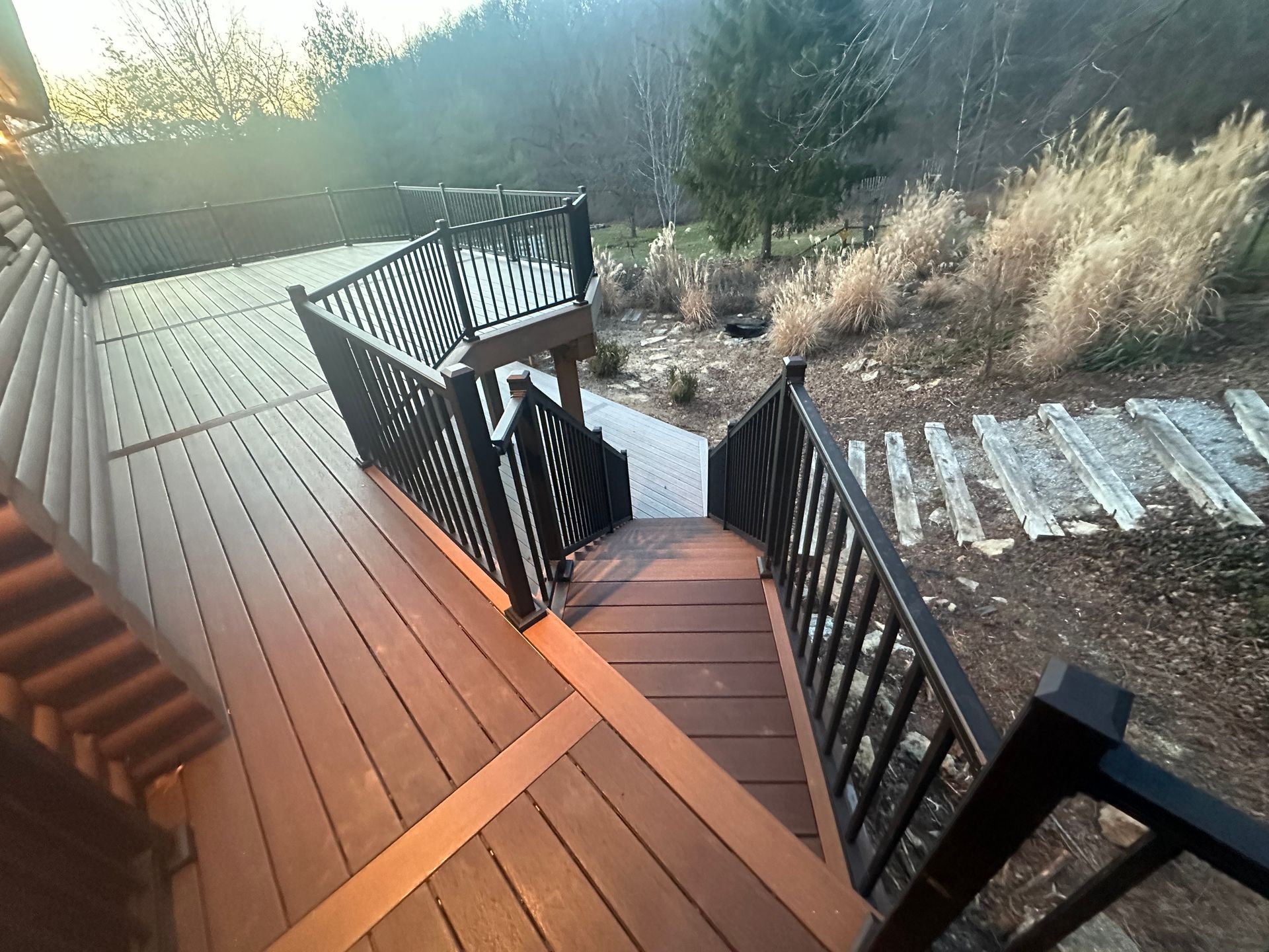 Wooden deck stairs with black railings leading down to a garden path in bright sunlight