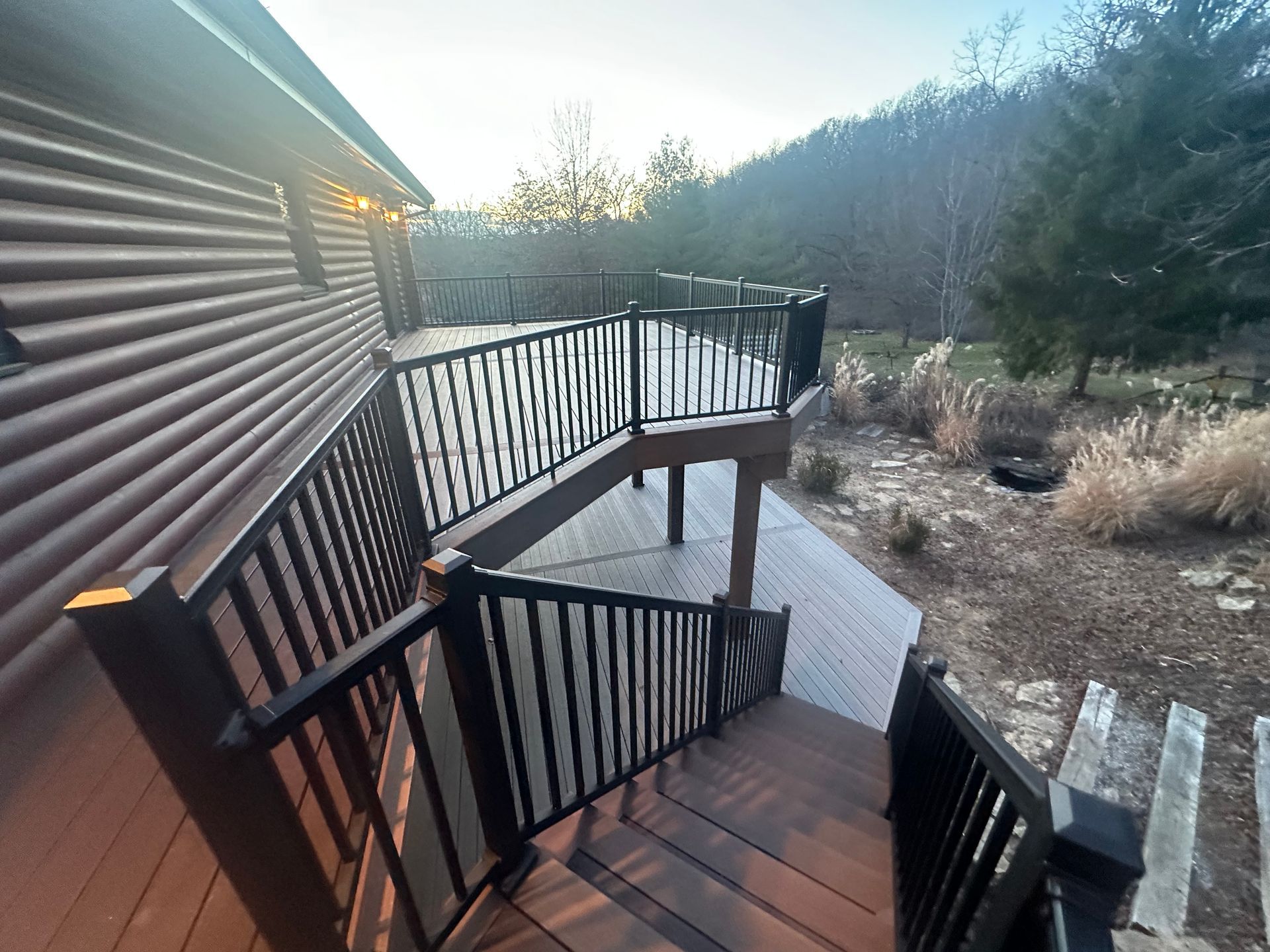 Wooden deck with black railings overlooking a grassy yard and wooded hillside at sunset