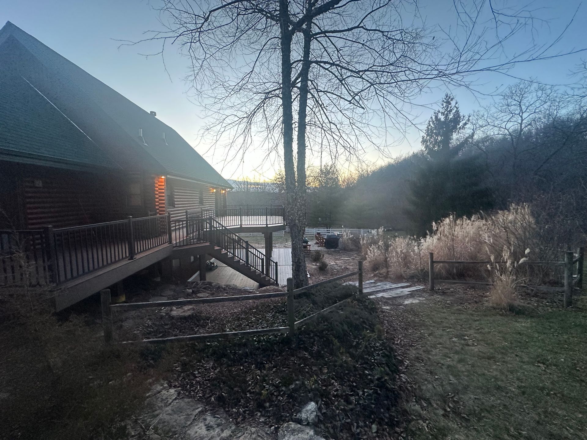 Dusk view of a wooden cabin by a stream, with leafless trees and a glowing porch light.