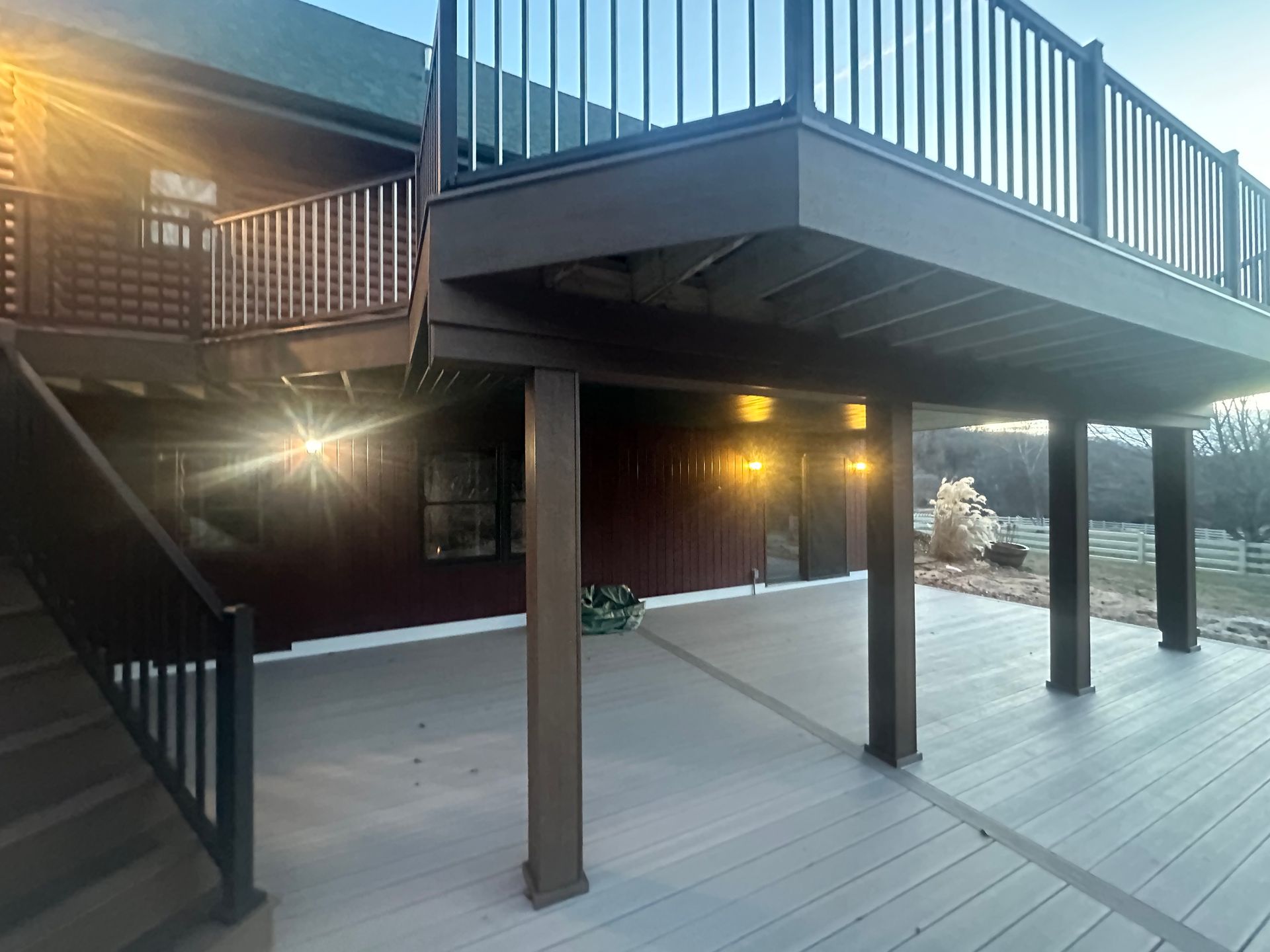 Snowy deck under an elevated balcony with dark wooden posts and stairs at dusk