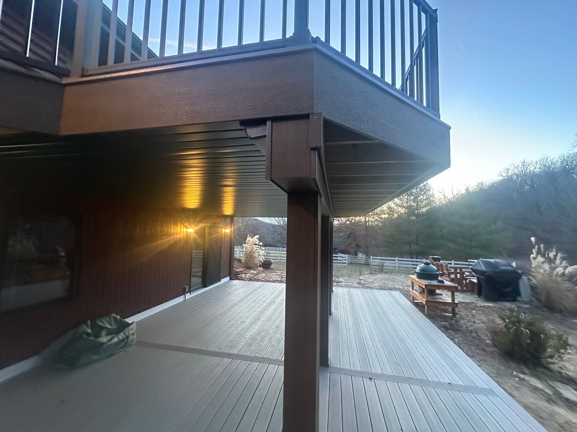 Wooden deck under a house overhang at sunset, with snow-covered ground and parked vehicles nearby
