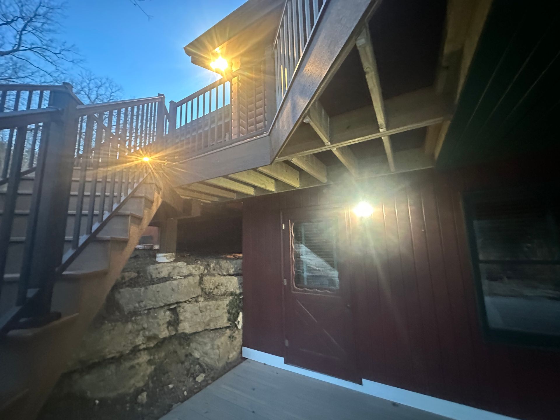 Back deck stairs and red doors beside a stone wall, with sunlight shining through the railing.